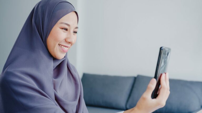 young asia muslim businesswoman using smart phone talk friend by videochat brainstorm online meeting while remotely work from home living room 1024x576 1